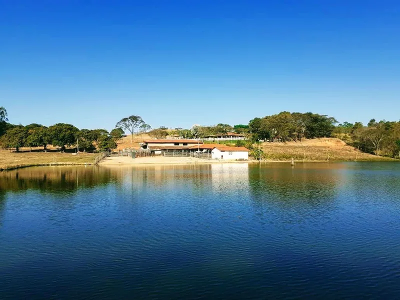 Clínica de Recuperação em Itaúna - MG Lago de água escura refletindo o céu azul, com construções térreas e pasto na margem oposta